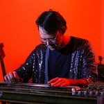 A colour photo shows artist Wayne Patrick Garret looking down and playing a pedal stell guitar. They are wearing a shimmering purple jacket and white glasses. A bright red background is behind them. 