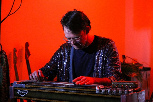 A colour photo shows artist Wayne Patrick Garret looking down and playing a pedal stell guitar. They are wearing a shimmering purple jacket and white glasses. A bright red background is behind them. 