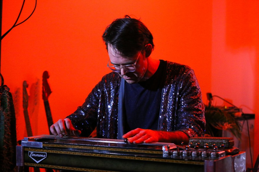 A colour photo shows artist Wayne Patrick Garret looking down and playing a pedal stell guitar. They are wearing a shimmering purple jacket and white glasses. A bright red background is behind them. 