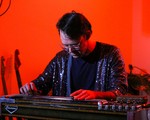 A colour photo shows artist Wayne Patrick Garret looking down and playing a pedal stell guitar. They are wearing a shimmering purple jacket and white glasses. A bright red background is behind them. 