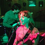 A colour photo featuring two members of Window Lamp Corp performing onstage, washed in pink and green light. 