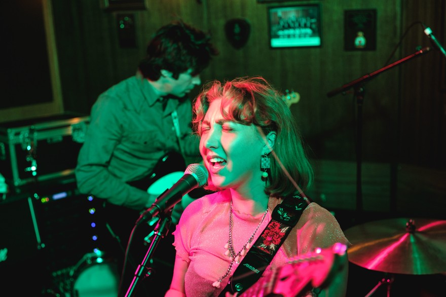 A colour photo featuring two members of Window Lamp Corp performing onstage, washed in pink and green light. 
