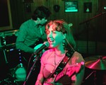 A colour photo featuring two members of Window Lamp Corp performing onstage, washed in pink and green light. 