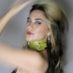 A blurry colour photo of Yolande Laroche posed with an arm on their head against a grey background. They're wearing a green leather accessory around thier night.  