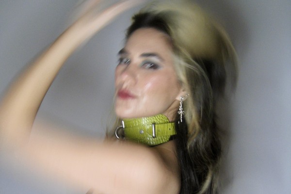 A blurry colour photo of Yolande Laroche posed with an arm on their head against a grey background. They're wearing a green leather accessory around thier night.  
