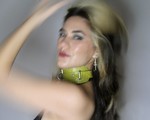 A blurry colour photo of Yolande Laroche posed with an arm on their head against a grey background. They're wearing a green leather accessory around thier night.  