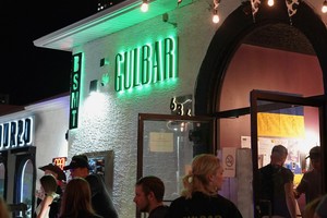 Nighttime scene outside Gulbar, where people gather under green neon signs and strings of cow‑patterned flags. The white building’s arched doorway glows warmly as guests stand, chat, and sit at the outdoor counter, creating a lively, social atmosphere.