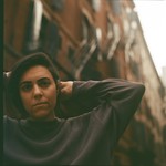 Sarah Tudzin stands in an urban alleyway lined with tall brick buildings and fire escapes, their hands lifted behind their head. Soft, warm light falls across the scene, highlighting the contrast between the dark sweatshirt they’re wearing and the textured architecture surrounding them.