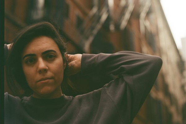Sarah Tudzin stands in an urban alleyway lined with tall brick buildings and fire escapes, their hands lifted behind their head. Soft, warm light falls across the scene, highlighting the contrast between the dark sweatshirt they’re wearing and the textured architecture surrounding them.