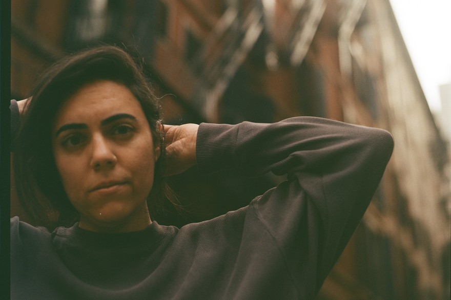 Sarah Tudzin stands in an urban alleyway lined with tall brick buildings and fire escapes, their hands lifted behind their head. Soft, warm light falls across the scene, highlighting the contrast between the dark sweatshirt they’re wearing and the textured architecture surrounding them.