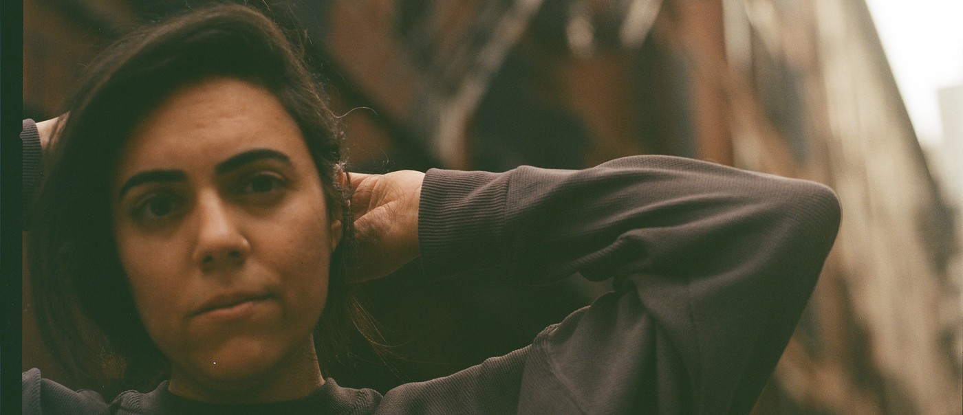 Sarah Tudzin stands in an urban alleyway lined with tall brick buildings and fire escapes, their hands lifted behind their head. Soft, warm light falls across the scene, highlighting the contrast between the dark sweatshirt they’re wearing and the textured architecture surrounding them.