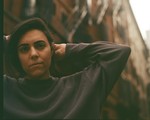 Sarah Tudzin stands in an urban alleyway lined with tall brick buildings and fire escapes, their hands lifted behind their head. Soft, warm light falls across the scene, highlighting the contrast between the dark sweatshirt they’re wearing and the textured architecture surrounding them.