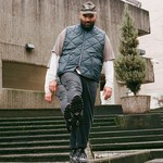 Justin R. Cruz Gallego stands on concrete steps outside a modern building, wearing a gray cap, blue quilted vest, and dark pants. One foot is lifted toward the camera, showing the tread of their shoe, while potted plants and textured concrete walls frame the urban scene.