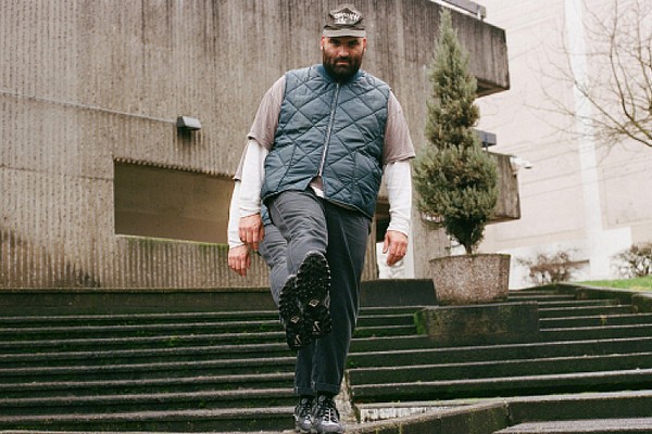 Justin R. Cruz Gallego stands on concrete steps outside a modern building, wearing a gray cap, blue quilted vest, and dark pants. One foot is lifted toward the camera, showing the tread of their shoe, while potted plants and textured concrete walls frame the urban scene.