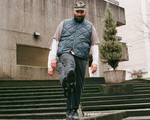 Justin R. Cruz Gallego stands on concrete steps outside a modern building, wearing a gray cap, blue quilted vest, and dark pants. One foot is lifted toward the camera, showing the tread of their shoe, while potted plants and textured concrete walls frame the urban scene.