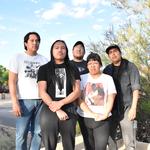 Five members of Weedrat stand together on a sunny residential street, framed by trees, bushes, and houses in the background. They wear casual graphic T‑shirts and jeans, posing in a relaxed group formation on the sidewalk.
