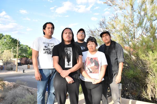 Five members of Weedrat stand together on a sunny residential street, framed by trees, bushes, and houses in the background. They wear casual graphic T‑shirts and jeans, posing in a relaxed group formation on the sidewalk.