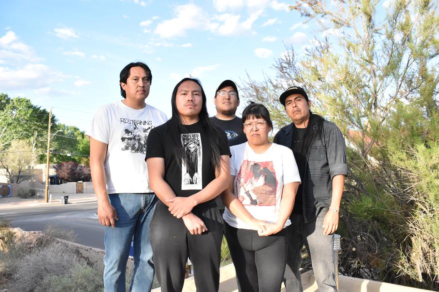 Five members of Weedrat stand together on a sunny residential street, framed by trees, bushes, and houses in the background. They wear casual graphic T‑shirts and jeans, posing in a relaxed group formation on the sidewalk.