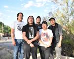 Five members of Weedrat stand together on a sunny residential street, framed by trees, bushes, and houses in the background. They wear casual graphic T‑shirts and jeans, posing in a relaxed group formation on the sidewalk.