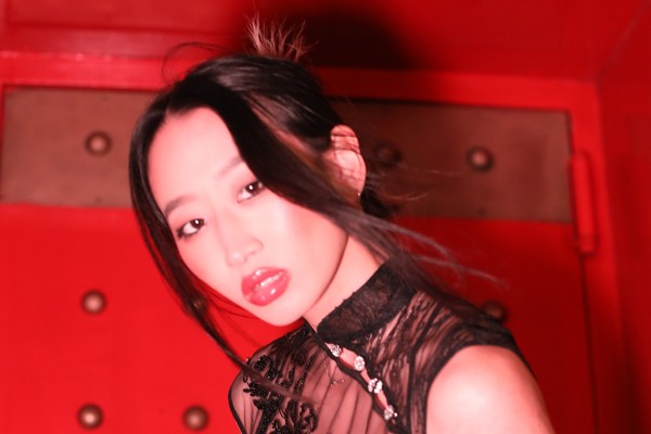Lil Mariko leans sideways against a vivid red, studded wall, wearing a sheer black garment embroidered with intricate patterns and beadwork. Dramatic lighting highlights the glossy shine of their lips and the delicate detailing of the outfit, creating a bold, high‑contrast portrait.