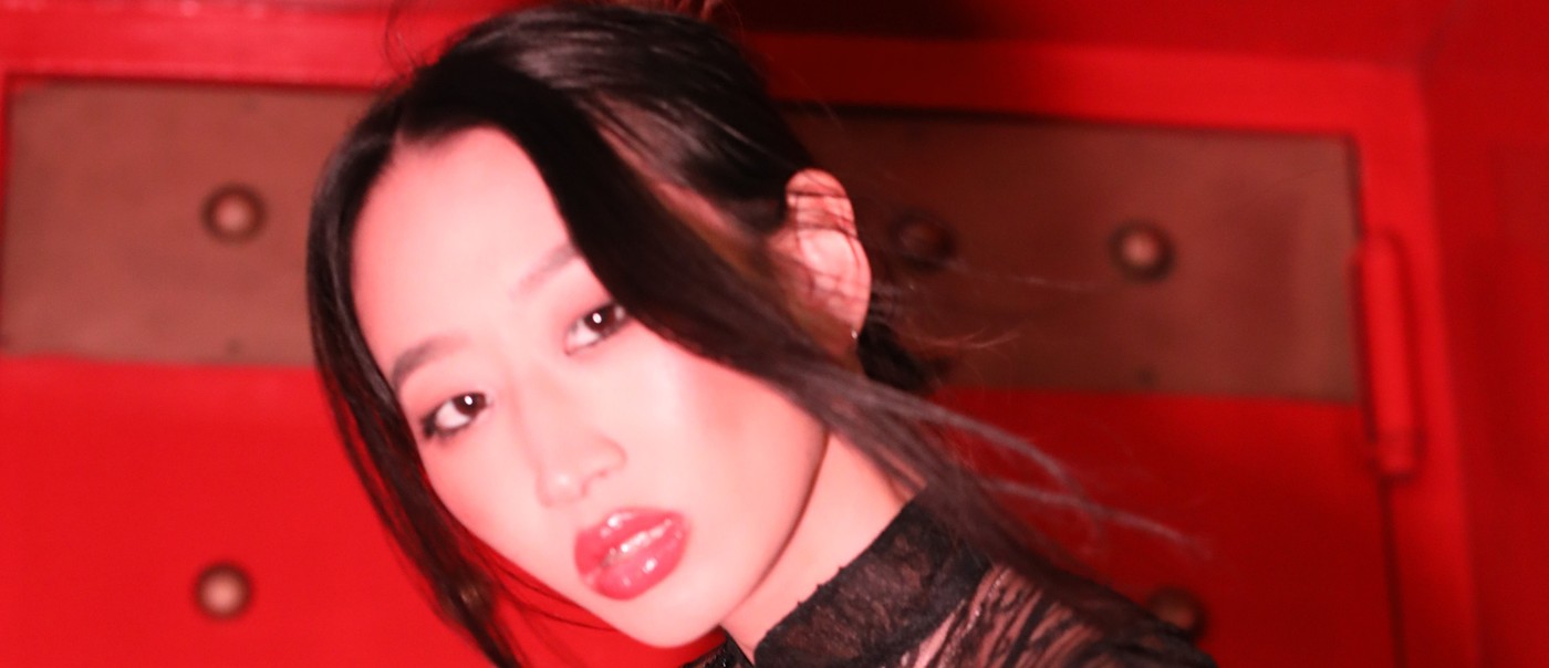 Lil Mariko leans sideways against a vivid red, studded wall, wearing a sheer black garment embroidered with intricate patterns and beadwork. Dramatic lighting highlights the glossy shine of their lips and the delicate detailing of the outfit, creating a bold, high‑contrast portrait.