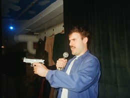 Comedian Ben Sosa‑Wright stands indoors in front of a dark green curtain, holding a microphone in one hand and a silver prop gun in the other. They wear a blue jacket over a light shirt, with hanging clothes and exposed ceiling pipes visible behind them, creating a playful, theatrical backstage vibe.