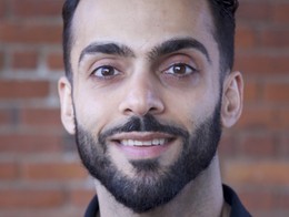 Portrait of comedian Mikey Dubs standing against a reddish‑brown brick wall, wearing a black collared shirt. Soft natural light highlights his short dark hair and neatly groomed beard, creating a clean, professional headshot.