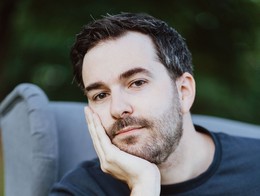 Comedian John Cullen sits outdoors in a gray cushioned chair, resting his head on one hand with his elbow propped on the armrest. He wears a dark long‑sleeved shirt with a small colorful graphic, framed by softly blurred greenery in the background.