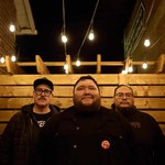 Three members of Iron Tusk stand together at night in a narrow outdoor space lit by hanging bulbs. They’re framed by brick walls and a wooden fence structure, their faces warmly illuminated as they pose in caps, glasses, hoodies, and jackets for a moody, urban group portrait.