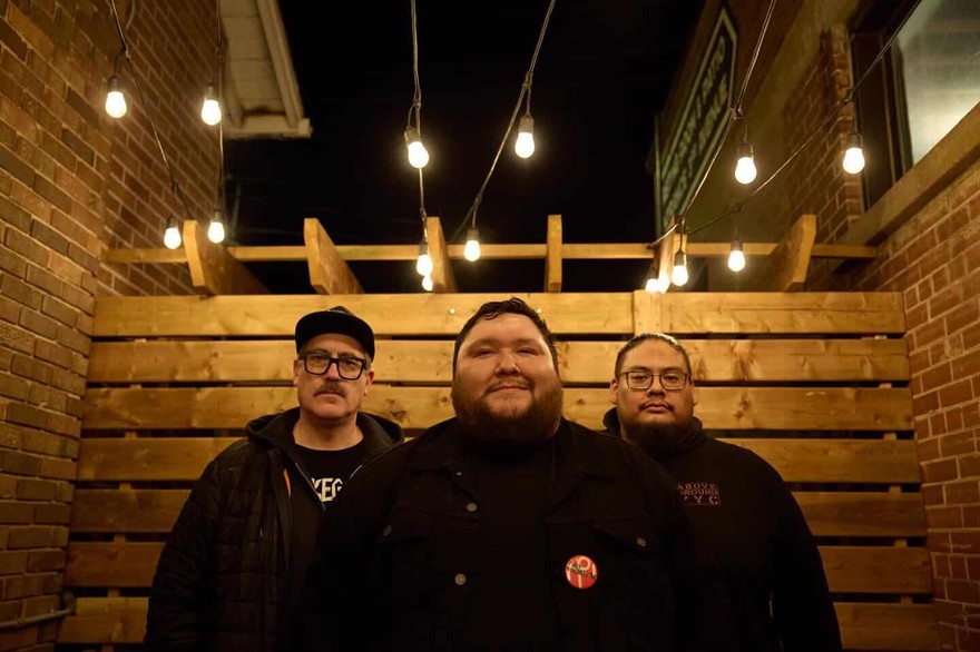 Three members of Iron Tusk stand together at night in a narrow outdoor space lit by hanging bulbs. They’re framed by brick walls and a wooden fence structure, their faces warmly illuminated as they pose in caps, glasses, hoodies, and jackets for a moody, urban group portrait.