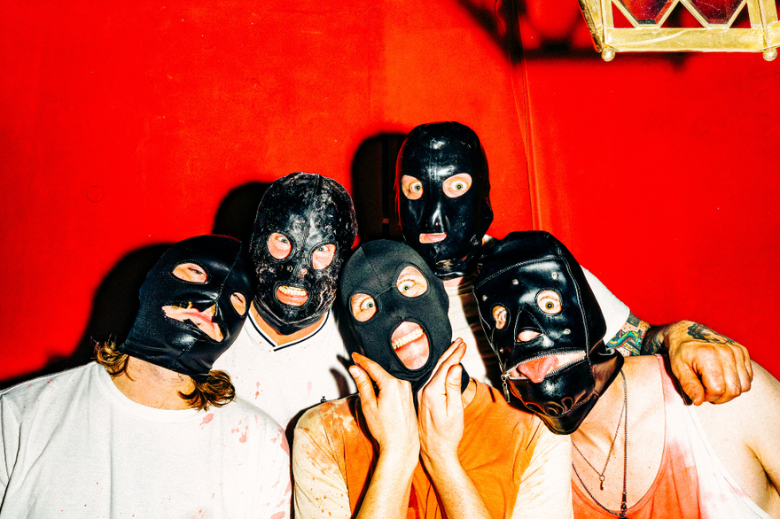 Five members of Tear Dungeon stand close together in front of a vivid red backdrop, each wearing a black mask that obscures most of their face. They’re dressed in casual shirts—some splattered or stained—and lit brightly against the intense background, creating a stark, unsettling group portrait.
