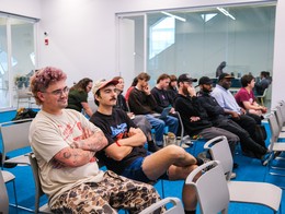 Attendees sit in rows of chairs at the Sled Island 2025 Conference inside a modern room with blue flooring and large glass walls. People face forward toward an unseen speaker, some with arms crossed, others listening intently, framed by the clean lines and reflective surfaces of the contemporary space.