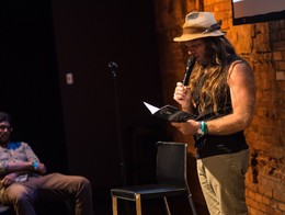 Thor Harris stands on a small indoor stage at the Sled Island 2015 Conference, reading from a booklet while holding a microphone. He wears a hat, sleeveless shirt, and light pants, with a chair beside him and a brick wall behind. Another attendee sits in the background, watching in the dim, intimate venue.