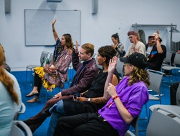 Attendees sit in a classroom‑style room at the Sled Island 2025 Conference, blue carpet beneath rows of gray chairs. Several people raise their hands to participate, facing a blank whiteboard at the front as they engage in a workshop‑like discussion.
