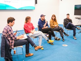 Five panelists sit in a row at the Sled Island 2025 Conference, each in a gray chair on blue carpet. One moderator on the left holds a microphone and notebook, while another panelist in a blue hoodie and cap speaks into a mic. The others listen or smile, framed by a large screen behind them displaying event text.