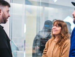 Three people stand in conversation inside a bright, modern space with glass walls and minimal furniture for the Sled Island 2025 conference. One person in a brown sweater faces two others—one in a blue hoodie and cap, the other dressed in dark clothing—as they talk casually near the partitioned area.