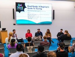 A panel of five speakers sits at the front of a conference room facing an audience. A large screen behind them displays the title “Sled Island Presents: Road Ready – A Beginner’s Guide to Touring,” along with the moderator and panelists’ names. Attendees sit in rows of gray chairs on blue carpet, listening as the discussion takes place. A podium with a laptop stands to the left of the panel.