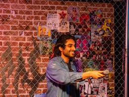 Comedian Amar Singh stands on a small stage holding a microphone, framed by a chain‑link fence and a brick wall plastered with bright, overlapping posters. He wears a distressed denim shirt and dark patched pants, lit by warm stage lighting with a stool nearby, capturing an intimate, DIY performance vibe.