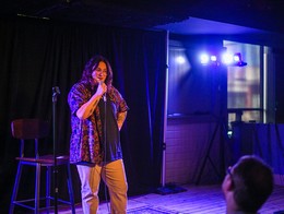Gabby Rose performs on a small stage at Sled Island 2024, standing in front of a black curtain and lit by blue and purple stage lights. She holds a microphone as audience members watch from the foreground, creating an intimate, low‑lit live performance atmosphere.