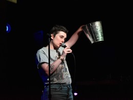 Comedian Sophie Haze performs on a small stage, holding a microphone in one hand and a silver bucket in the other.