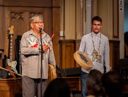 Two presenters stand on a wooden‑paneled stage during the 2025 Elder Blessing, Elder Aapi’ohkitopi (Grey Horse Rider) speaks into a microphone while holding a decorated stick, the other holding a hand drum painted with an animal design. Both wear patterned clothing and beadwork, framed by guitars and drums in the background, creating a warm, ceremonial atmosphere.