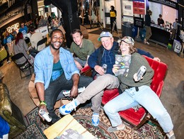 Four people relax together at Sled Village 2025, sitting on a red couch and nearby chairs with canned drinks in hand. One person holds a Sled Island program guide as others smile toward the camera. Behind them, attendees mingle near tables and a counter, creating a warm, social festival atmosphere in an industrial‑style space.
