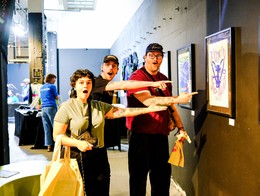 Three people stand in at the Sled Island Poster show pointing excitedly at a framed artwork on the wall. The piece features bold stylized text and a purple-toned graphic. Additional framed works hang along the gallery wall, and other visitors browse displays in the brightly lit space behind them.