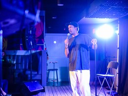 Comedian Faris Hytiaa performs on a stage at Sled Island 2024, holding a microphone and bathed in blue lighting.