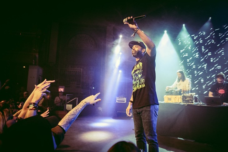 The front person of the hip-hop group clipping stands center stage under moody blue and purple lighting, holding a microphone aloft toward an enthusiastic crowd at The Palace Theatre during Sled Island 2026. He wears a black baseball cap, a black T-shirt with neon green and yellow text reading “STANDARD CALIFORNIA,” and jeans. Audience members reach out with raised hands and tattooed arms toward him. Behind the performer, two DJs operate turntables and laptops on a table, one in a white shirt and the other in a dark shirt with a red sunburst graphic. A large LED backdrop glows with abstract light patterns, adding energy to the indoor concert venue’s lively atmosphere.