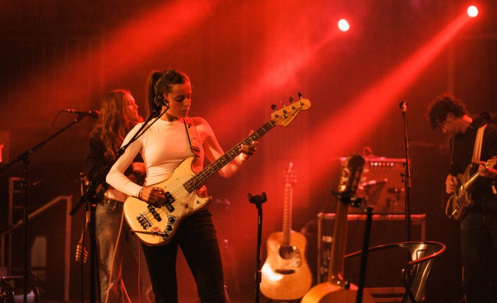 The band Black Country, New Road performs at The Palace Theatre under deep red stage lighting during Sled Island 2026. In the foreground, a musician wearing a white long‑sleeve top plays an electric bass guitar, focused on her instrument. Behind her, two other performers play electric guitars, partially illuminated by the warm glow. Microphones, amplifiers, and acoustic guitars rest on stands around them, adding to the intimate concert atmosphere.