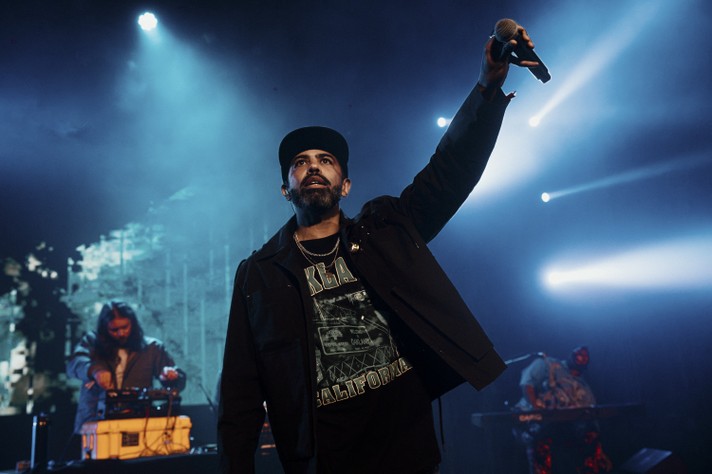 The frontperson of the hip-hop group clipping stands on stage under blue and white spotlights, holding a microphone high in one hand. He wears a black cap, dark jacket, and a T‑shirt that reads “Oakland California.” Behind him, two other musicians operate electronic equipment and keyboards, partially obscured by haze and beams of light. The scene captures the energy and atmosphere of a live music performance taking place at The Palace Theatre during Sled Island 2026.