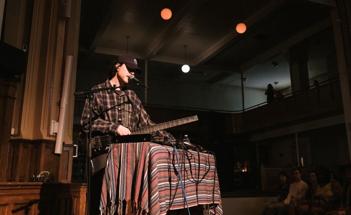 Solo musician HOMESHAKE performs indoors at a church venue during Sled Island 2026, standing behind a table draped with a striped, multicolored cloth. He wears a dark baseball cap and plaid shirt, singing into a microphone while playing a long, rectangular electric instrument. Audio cables and equipment are spread across the table. Warm lighting highlights the performer against the dim background, where a few audience members sit watching and others observe from a balcony above.