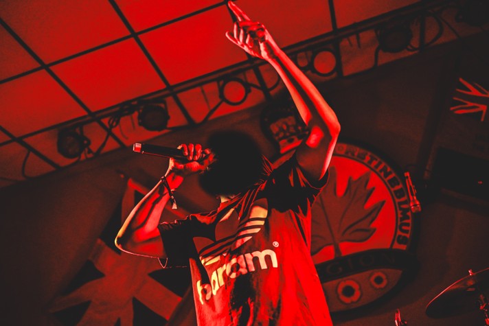Rapper billy woods stands on stage at the #1 Legion during Sled Island 2026 under intense red lighting, holding a microphone close to his mouth and raising one arm high with his index finger pointed upward. He wears a dark T‑shirt with a bold logo and the word “haram” printed across the front. Behind him, flags and a circular emblem featuring a maple leaf are visible, along with stage lights mounted on the ceiling. The vivid red tones create a dramatic, high‑energy atmosphere typical of a live music performance.