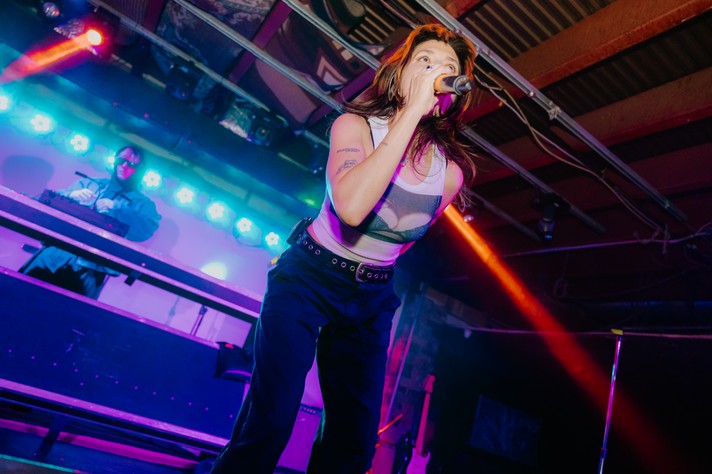 A performer from the band Sextile leans forward on stage at Dickens in Calgary, singing passionately into a microphone under vivid blue and red lighting during Sled Island 2026. She wears a sleeveless white top with a mesh design and dark pants with a studded belt, her hair glowing in colorful beams. Behind her, a DJ in sunglasses and a dark jacket operates equipment surrounded by bright LED lights, creating an electrifying atmosphere in the intimate venue.